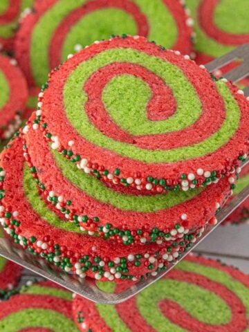 Bright red and green Christmas pinwheel cookies stacked on a metal spatula, coated with festive nonpareil sprinkles around the edges.
