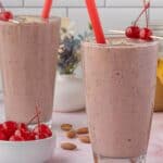Two tall glasses of cherry almond smoothie on a kitchen counter, each topped with cherries and served with red straws.