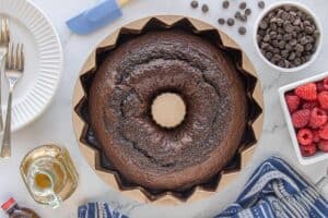 Freshly baked chocolate Bundt cake cooling in the pan on a kitchen counter, surrounded by ingredients, olive oil, chocolate chips and raspberries.