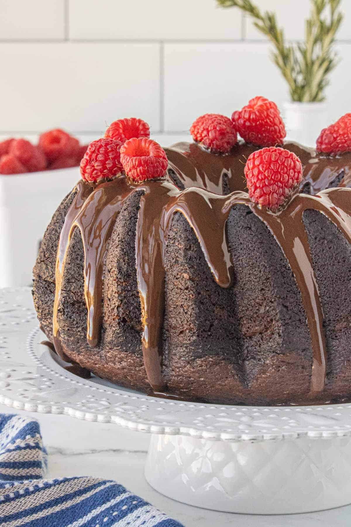 Chocolate olive oil bundt cake on a cake stand, drizzled with chocolate glaze and topped with fresh raspberries.