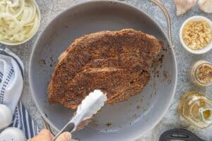 Beef roast being seared in a skillet with a dark spice rub forming a crust.
