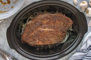 Seared beef roast placed on top of sliced onions inside a slow cooker.