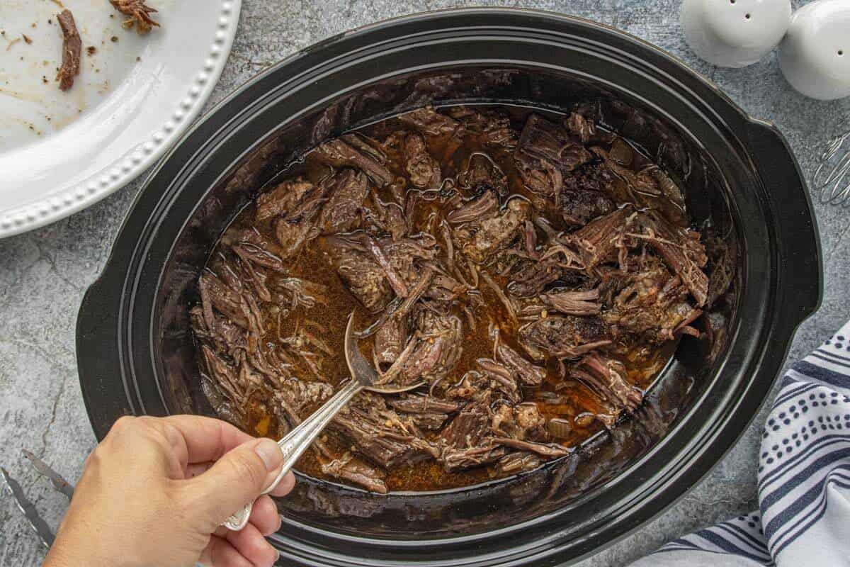 Fully cooked shredded beef inside the slow cooker with onions and rich cooking juices. A serving of shredded beef is being served with a large spoon.