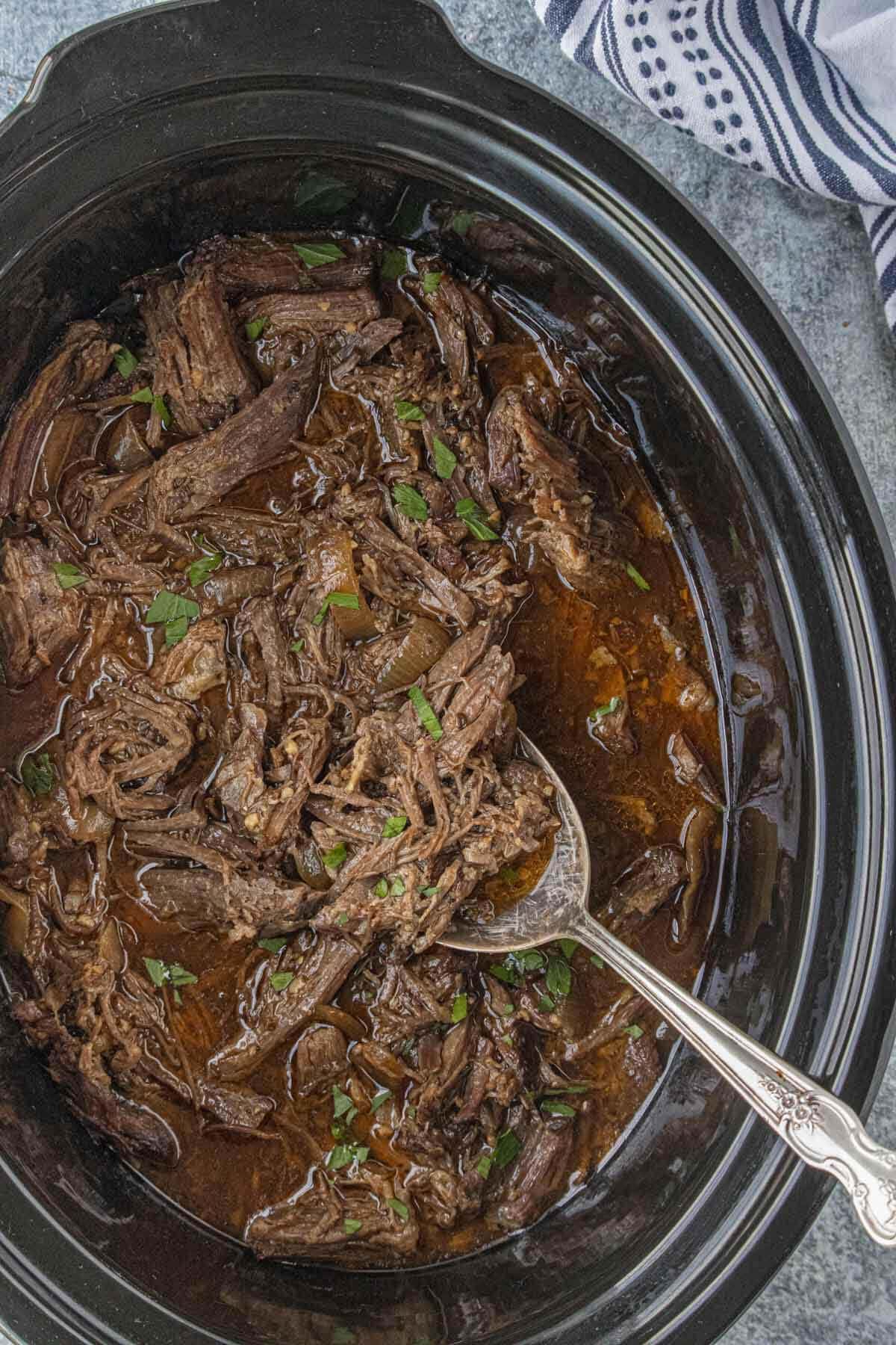 Slow cooker shredded beef shown from above with a spoon lifting the meat and broth.