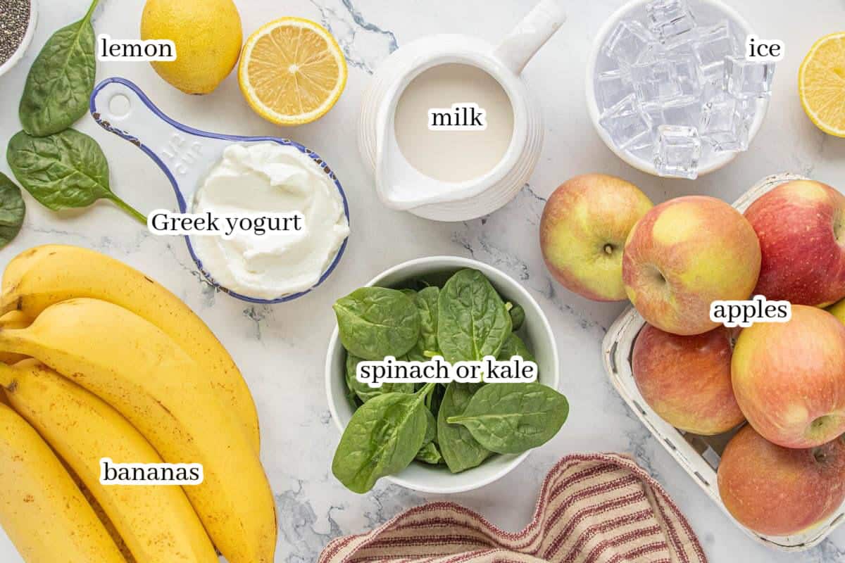 Overhead view of green smoothie ingredients on a marble surface, including bananas, apples, spinach or kale, Greek yogurt, milk, ice, and fresh lemon.