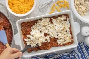 Creamy noodle mixture being spread over the beef layer in a greased baking dish.