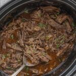Close-up of tender shredded beef garnished with minced parsley in a slow cooker, with a serving spoon.