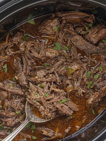 Close-up of tender shredded beef garnished with minced parsley in a slow cooker, with a serving spoon.