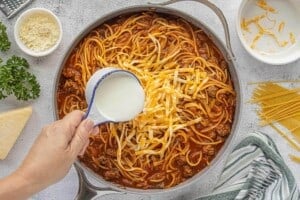 Shredded cheese and milk being added to pasta in a skillet to create a creamy sauce.