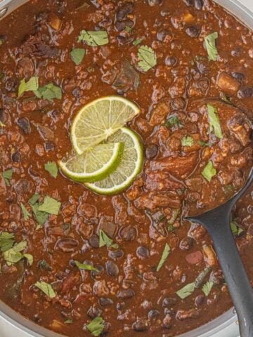 Overhead view of pot of chili with a ladle for serving, garnished with fresh herbs and lime slices.