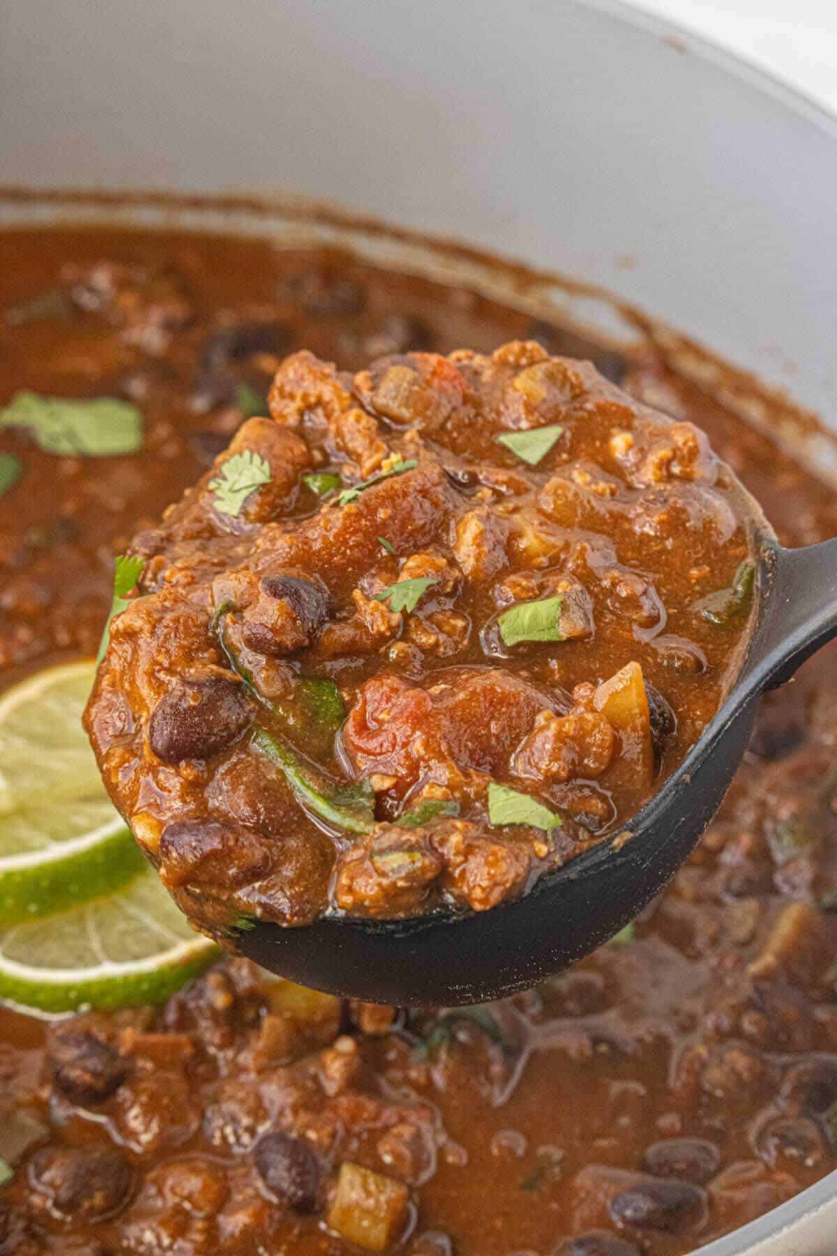 Close-up of a pot of chili with a ladle taking a big scoop. Showing tender ground turkey, black beans, zucchini, and peppers in a thick sauce.