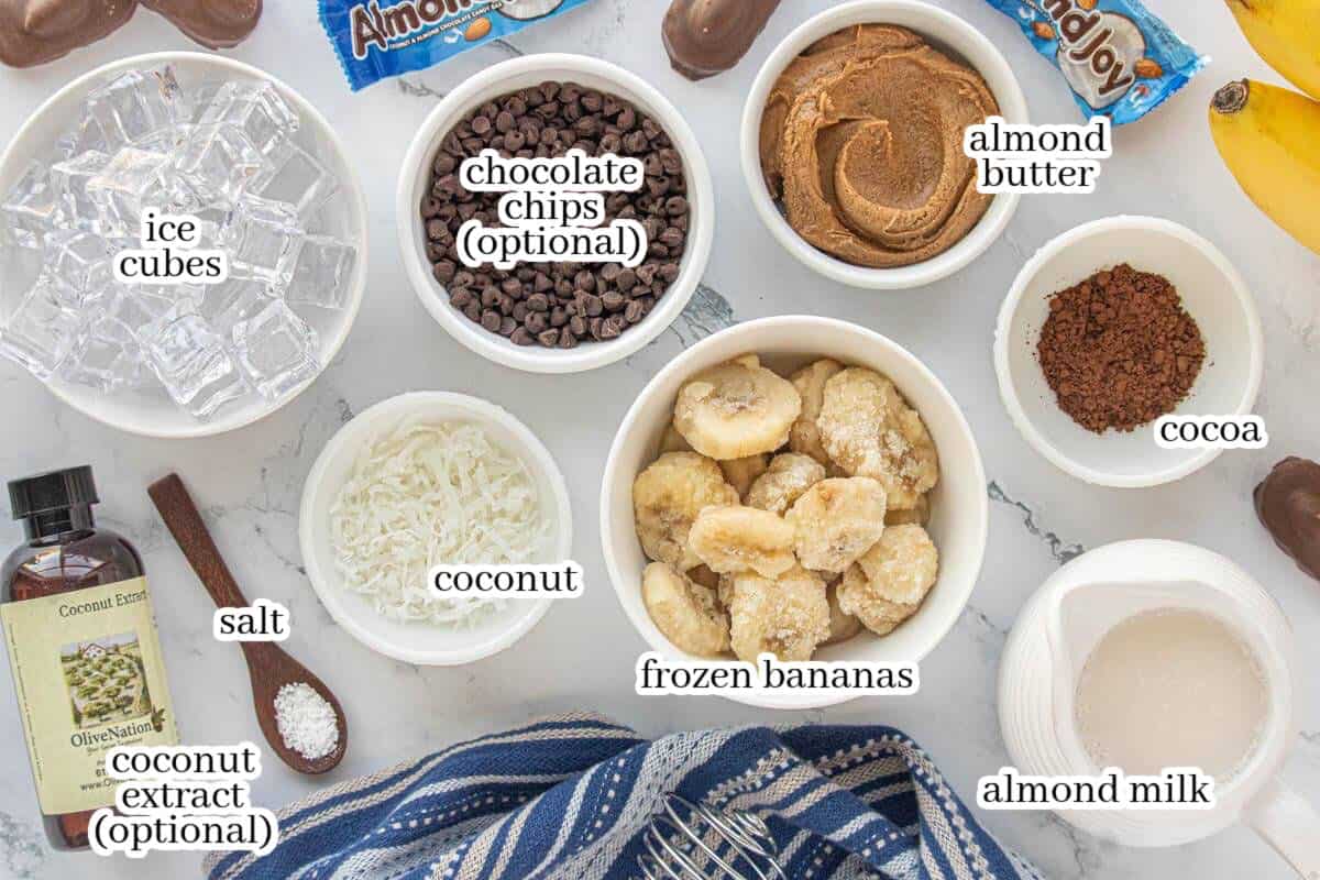 Ingredients for smoothie including frozen banana slices, almond butter, cocoa powder, shredded coconut, chocolate chips, almond milk, ice, and coconut extract.
