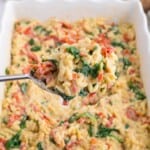 Spoon lifting creamy Boursin orzo pasta with spinach and sun-dried tomatoes from the baking dish.