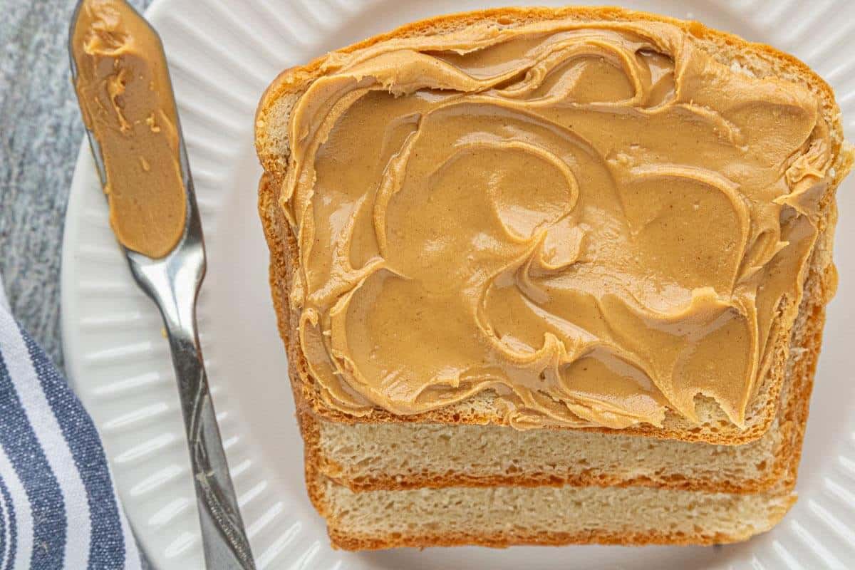 Slice of soft sandwich bread spread with peanut butter on a white plate.
