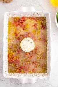 Uncooked orzo and sun-dried tomatoes layered in a white casserole dish with seasonings, stock and olive oil.