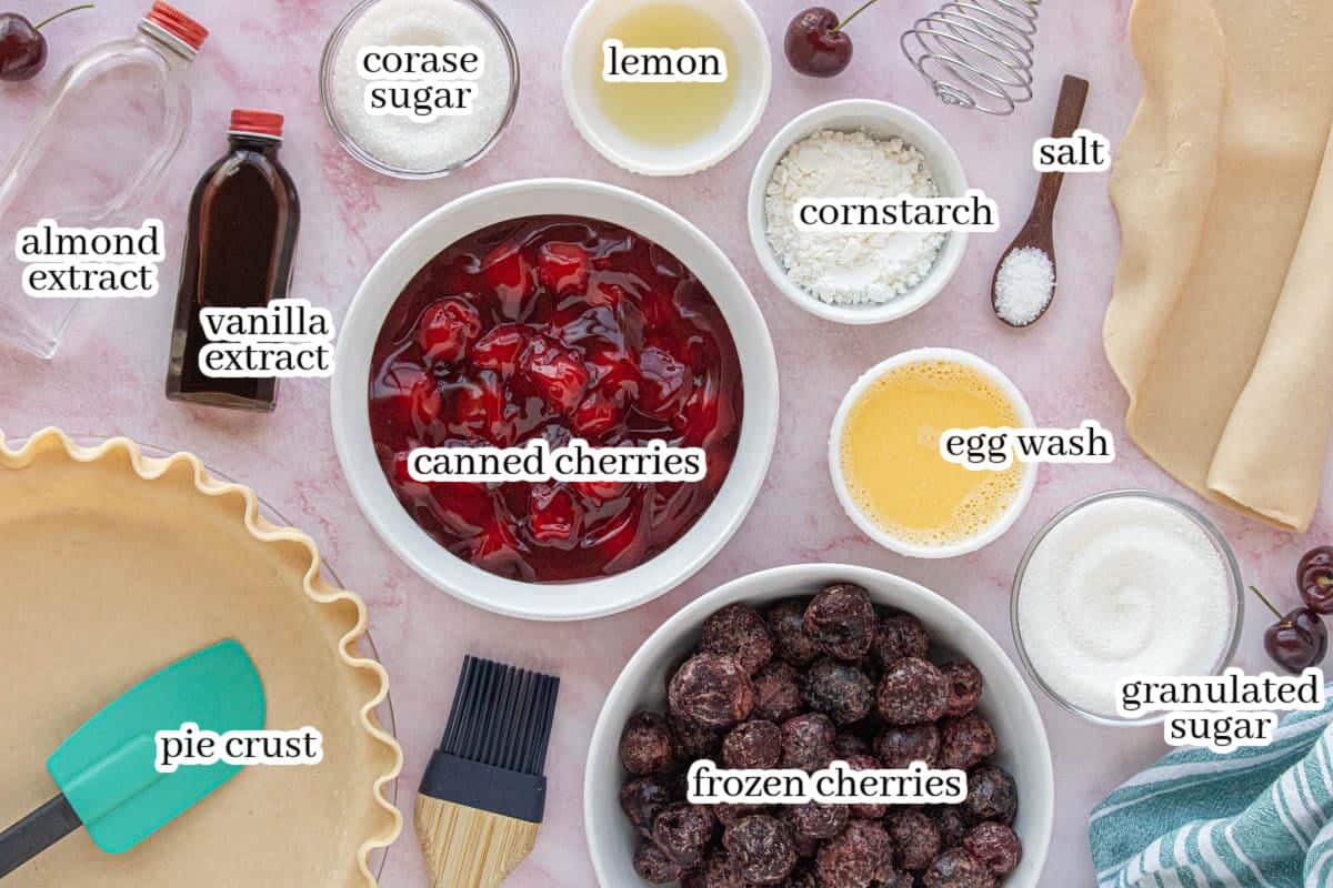Cherry pie ingredients laid out on a pink surface, including canned cherry pie filling, frozen cherries, pie crust, sugar, cornstarch, lemon juice, egg wash, vanilla extract, almond extract, and salt.