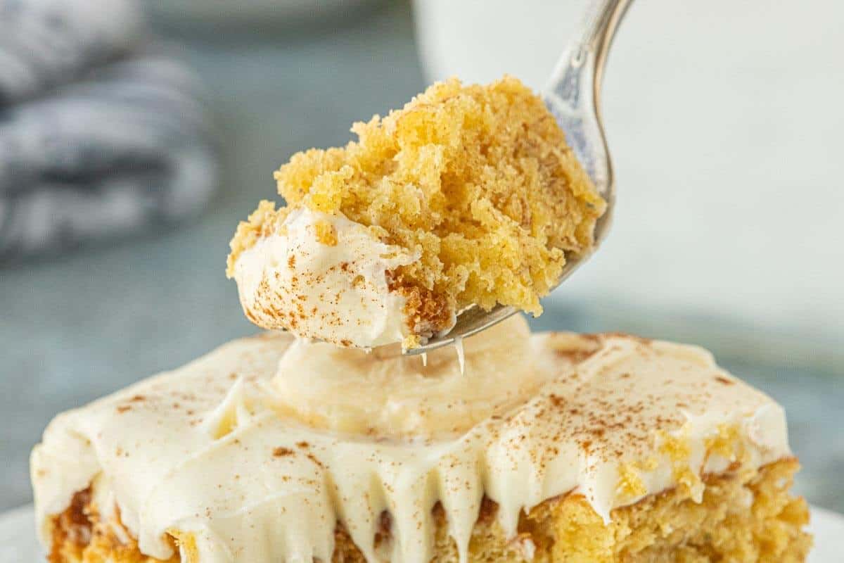 Slice of banana cake with cream cheese frosting on a fork above a slice of cake.