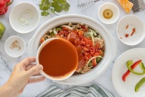 Hand pouring enchilada sauce over meatballs, peppers, and onions in a slow cooker.