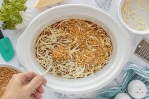 Chicken in the crockpot covered with sauce and seasoning before cooking, then topped with shredded mozzarella cheese and toasted breadcrumbs.