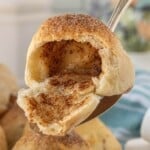 A golden brown baked roll broken open to reveal the hollow cinnamon sugar center, being held by a spoon.