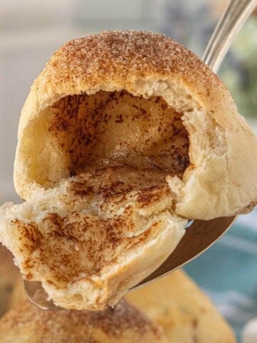 A golden brown baked roll broken open to reveal the hollow cinnamon sugar center, being held by a spoon.