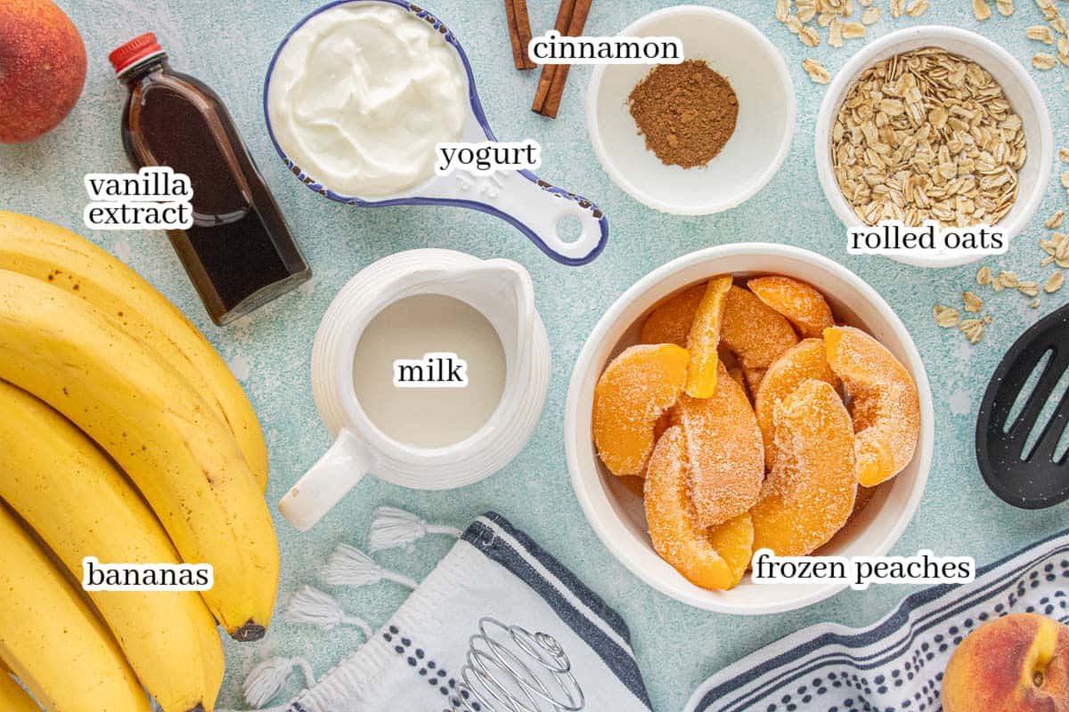 Ingredients for a peach smoothie including frozen peaches, banana, yogurt, milk, vanilla extract, cinnamon, and rolled oats arranged on a countertop.