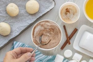 A bowl of cinnamon sugar and melted butter beside thawed dinner roll dough and marshmallows on a baking sheet.