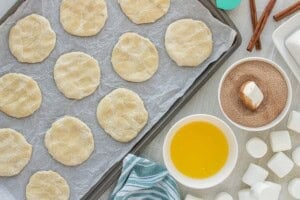 Flattened dough rounds arranged on baking sheet with melted butter nearby with a marshmallow being rolled in cinnamon sugar mixture.