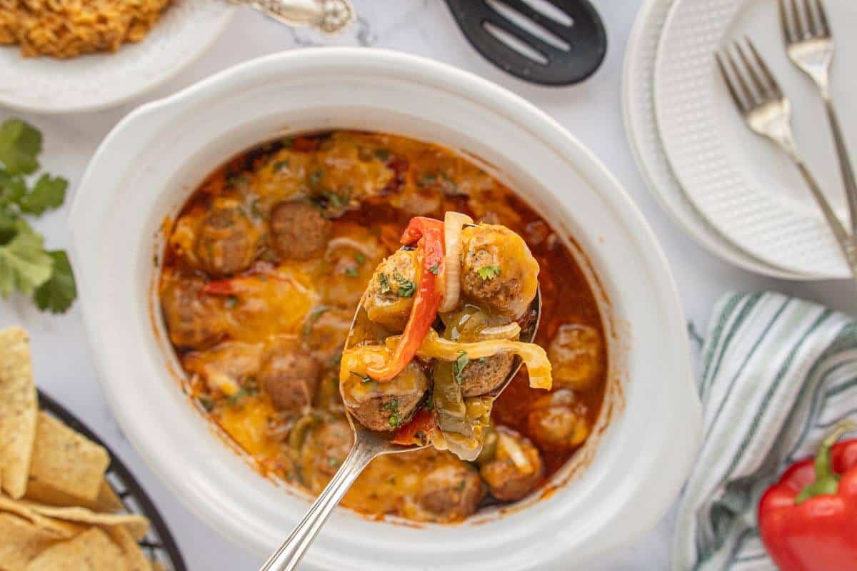Spoon lifting burrito meatballs, with peppers, onions, and melted cheese from a slow cooker filled with sauce.