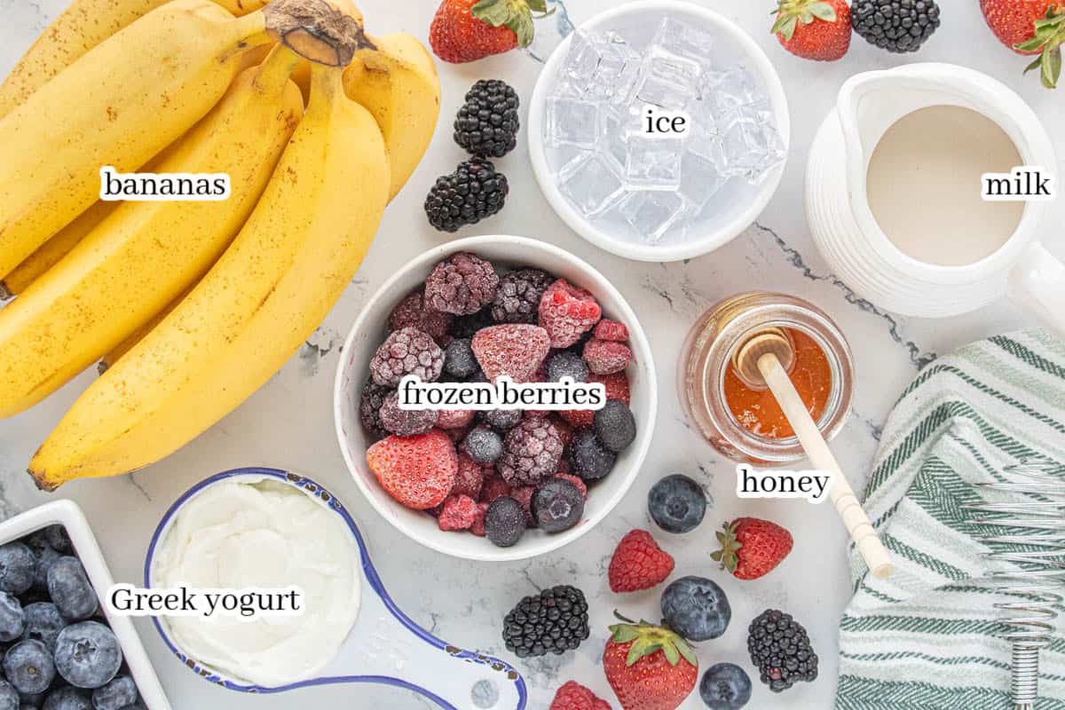 Ingredients for a berry banana smoothie including bananas, frozen mixed berries, Greek yogurt, milk, honey, ice, and fresh strawberries, blueberries, and blackberries on a countertop.