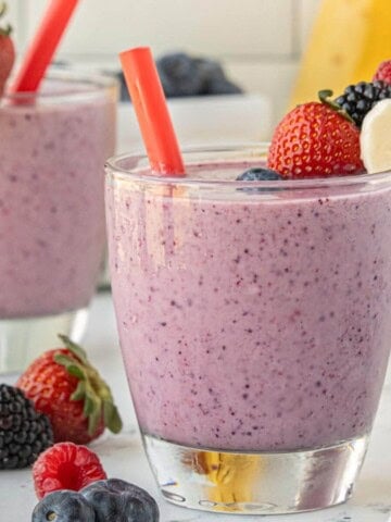 Creamy berry banana smoothie in a glass with a red straw, with fresh berries scattered around the base on a marble surface.