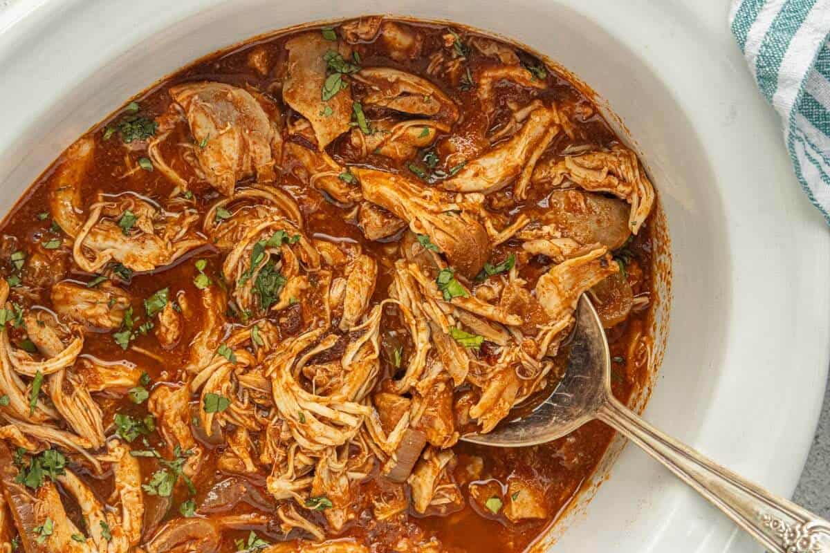 Close-up of saucy shredded tinga chicken simmering with onions in the crockpot.