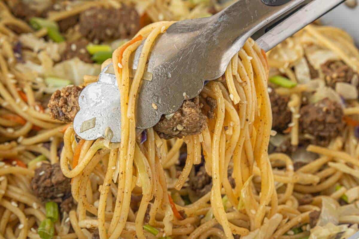 Tongs lifting a portion of ground beef chow mein noodles from a skillet showing texture and ingredients.