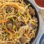 Overhead view of chow mein in a skillet with noodles, seasoned beef, and vegetables with red sauce on the side.