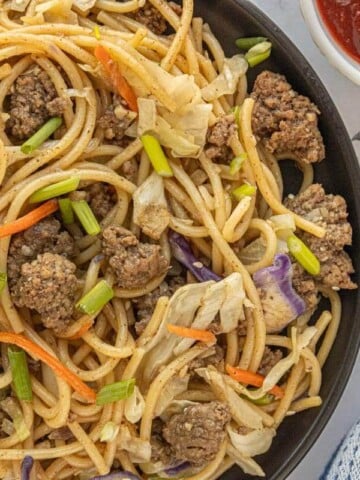 Overhead view of chow mein in a skillet with noodles, seasoned beef, and vegetables with red sauce on the side.