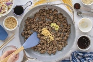 Ground beef cooking in a skillet with minced garlic and ginger being added.