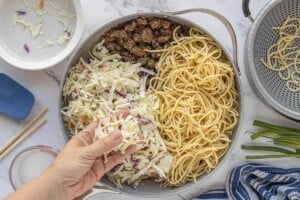Ground beef chow mein coming together in a skillet with noodles and coleslaw mix being added.