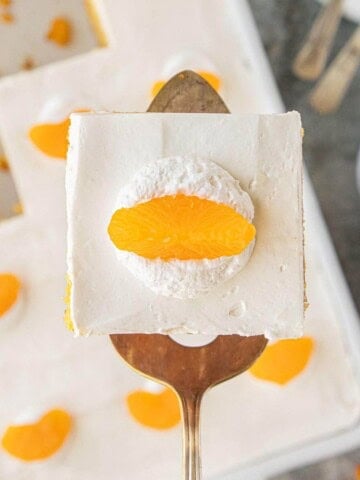 Slice of creamsicle poke cake on a serving spatula topped with whipped cream and a mandarin orange segment.