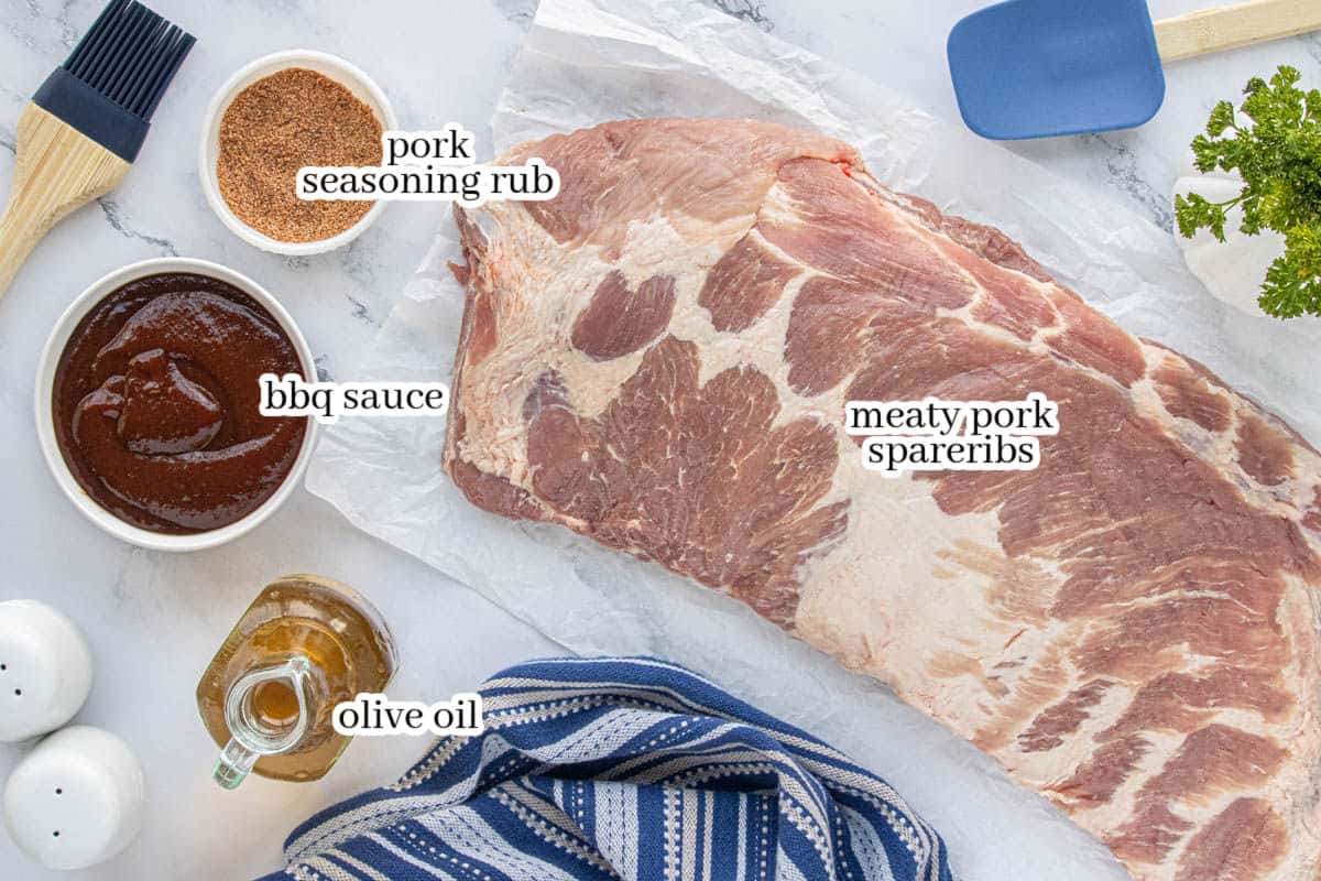 Raw pork spareribs on a countertop with olive oil, barbecue sauce, and seasoning rub arranged nearby for prep.