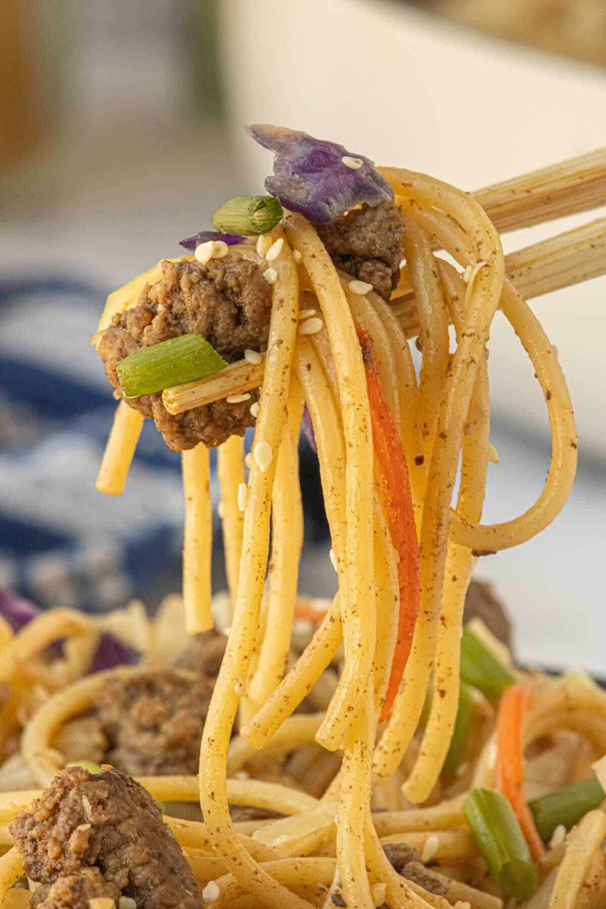 Close-up of spaghetti noodles lifted with chopsticks showing ground beef, cabbage, and sesame seeds.