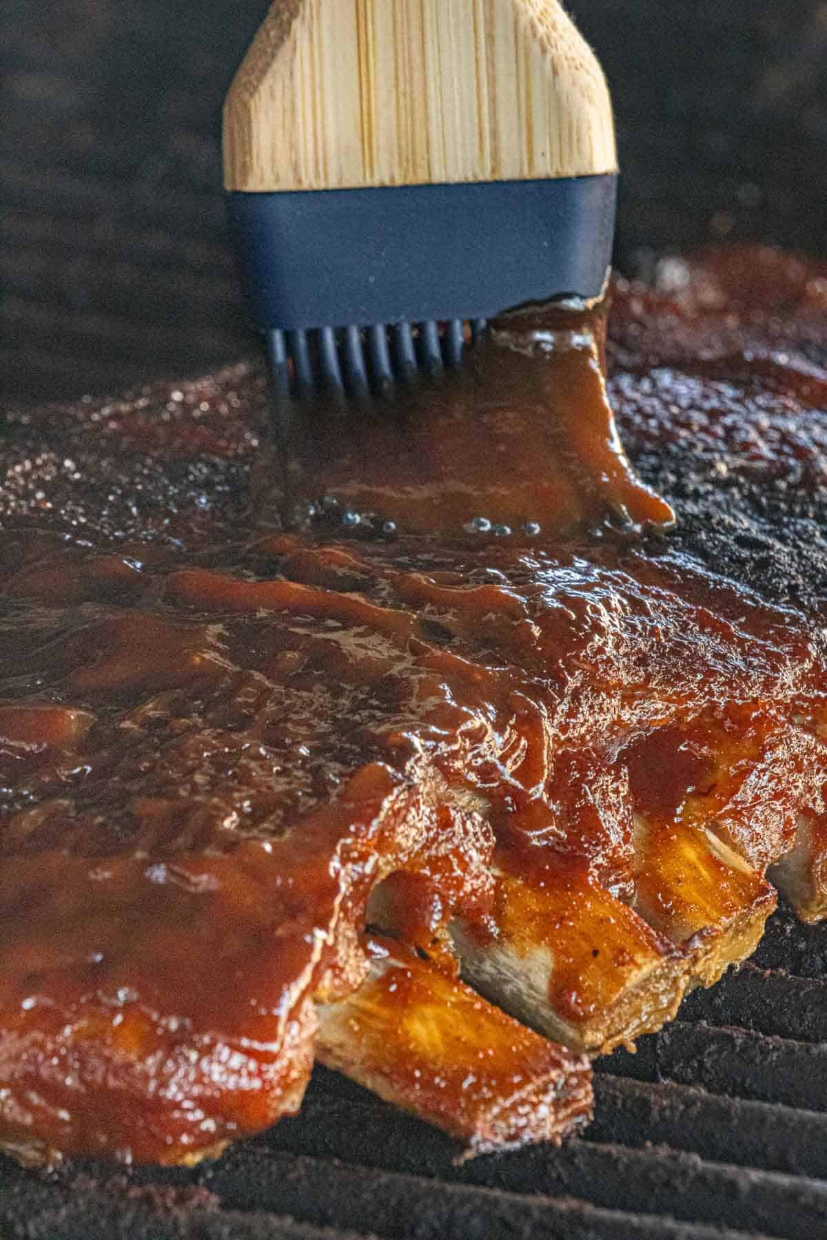 Barbecue ribs on a grill being brushed with sauce using a basting brush.