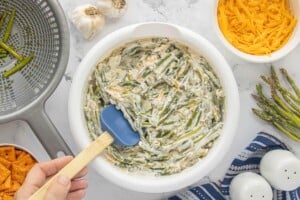 Asparagus folded into the sauce mixture in a white mixing bowl with a blue spatula.