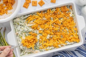 Creamy sparagus casserole in baking dish with cheesy cracker topping being spooned over the mixture prior to baking.
