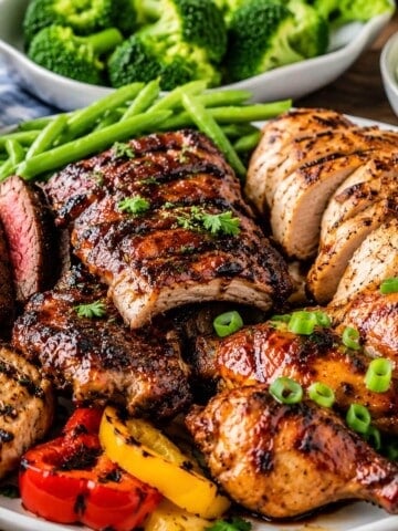 A platter filled with grilled chicken, ribs, chops and vegetables, garnished with chives. With green beans and broccoli in the background.