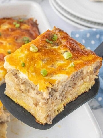 Slice of biscuits and gravy casserole with sausage, eggs, and melted cheddar cheese served from a baking dish on a black spatula.