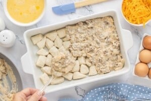 Creamy sausage gravy being spooned over unbaked biscuits in a casserole dish.