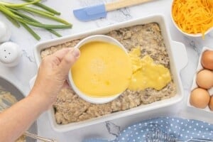 Beaten eggs being poured over the sausage gravy and biscuit layers in a casserole dish.