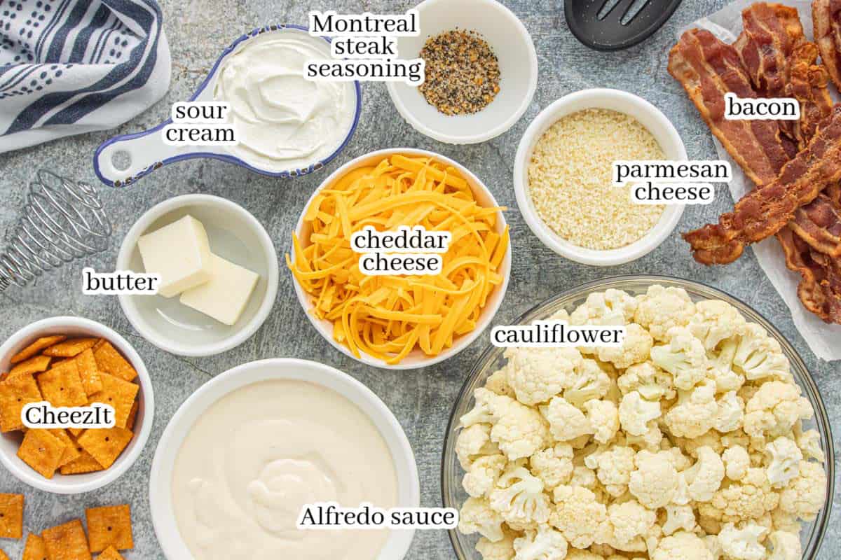 Ingredients for the casserole including cauliflower florets, cheddar cheese, Alfredo sauce, sour cream, Parmesan cheese, bacon, butter, Cheez-It crackers, and Montreal steak seasoning on a countertop.