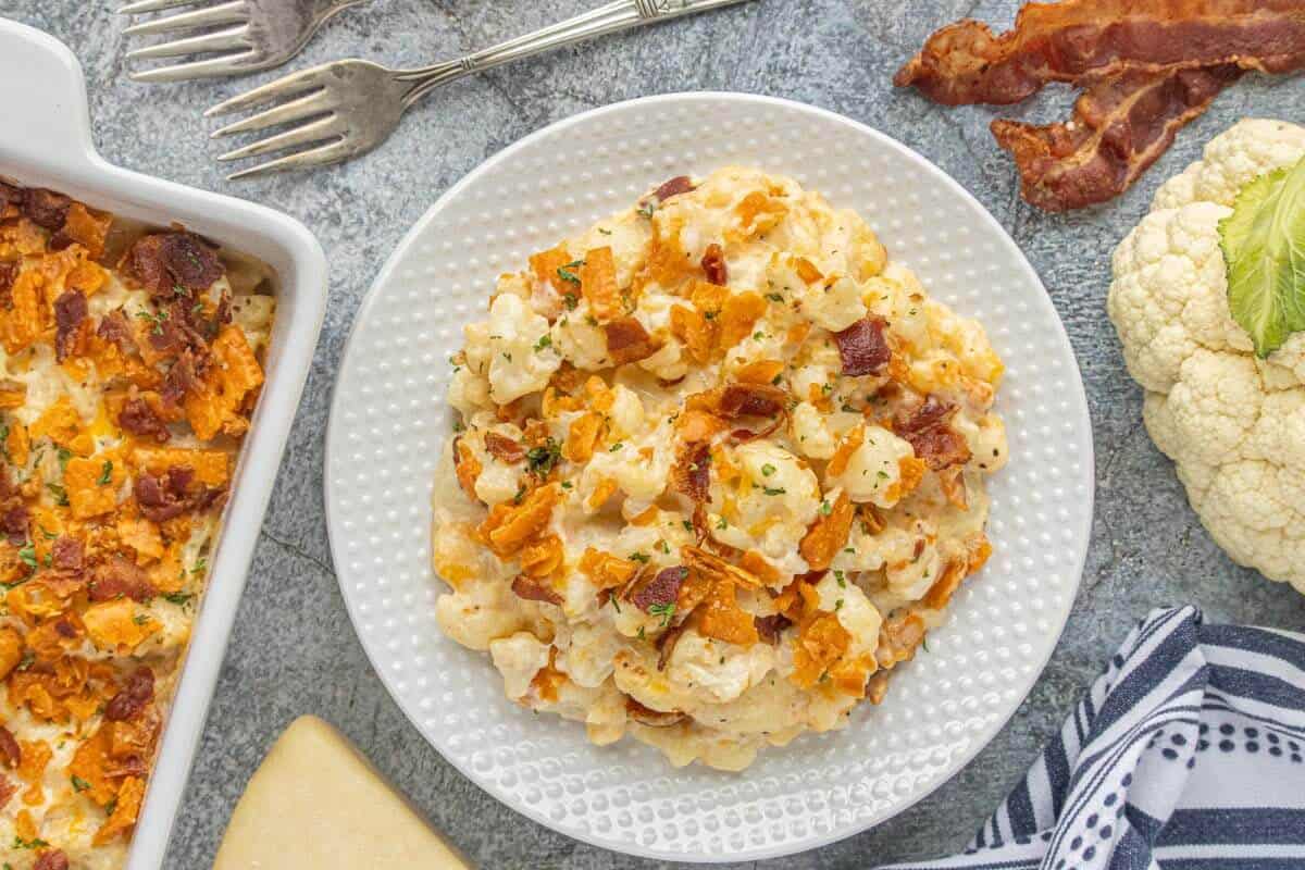 Cheesy cauliflower casserole topped with bacon and crunchy crackers served on a white plate beside a casserole dish.