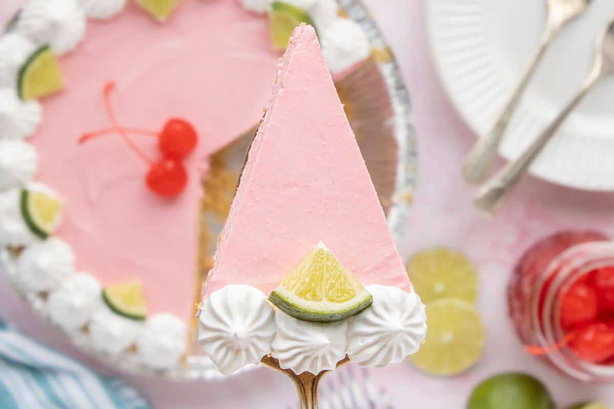 Close-up of a slice of No-Bake Cherry Limeade Pie topped with whipped cream and lime, showing the creamy pink filling and graham cracker crust.